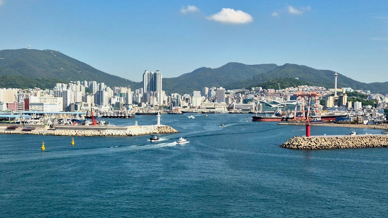 Busan port skyline with cranes, mountains, and boats entering the harbor on a clear day