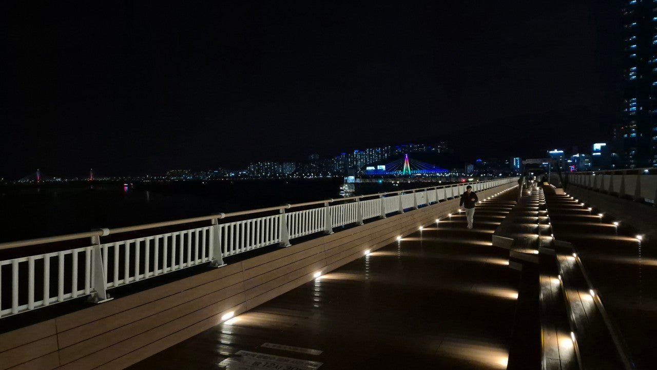 Illuminated wooden riverwalk deck at night near Gupo Station, Busan, with a lone walker
