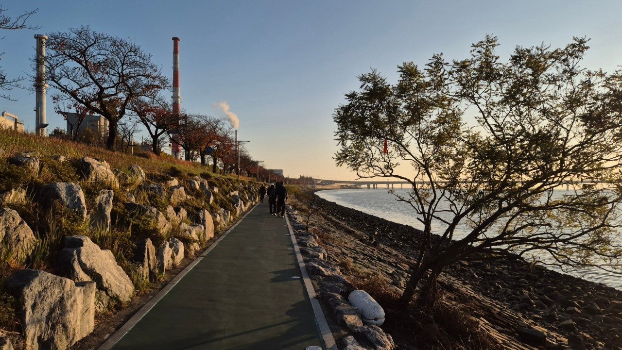 Riverside walking path along the Nakdong River near Jangnim, Busan, with walkers silhouetted at sunset and industrial chimneys in the background
